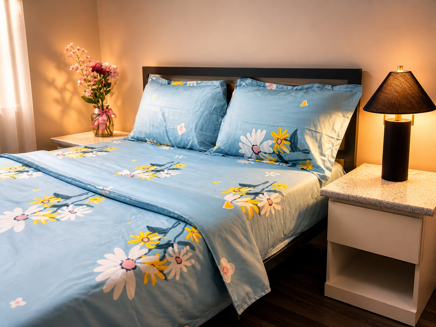Bed with floral bedding in a bedroom setting with a lamp and flowers on a nightstand.