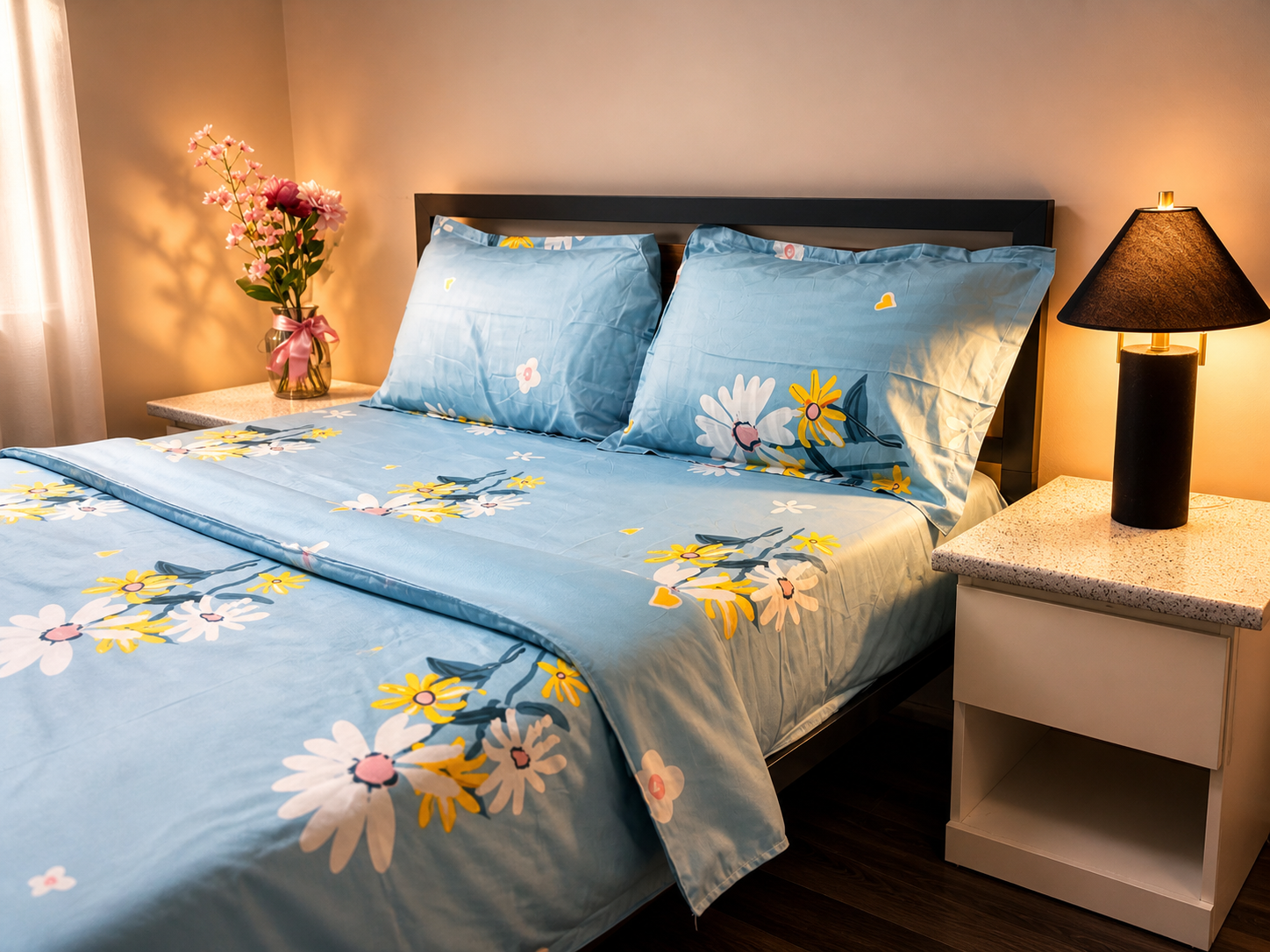 Bed with floral bedding in a bedroom setting with a lamp and flowers on a nightstand.